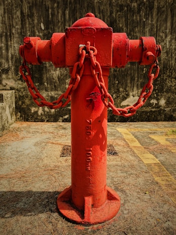 A bright red fire hydrant is prominently positioned in the center, with heavy chains attached to its sides. The hydrant is placed on a concrete surface marked with yellow lines and surrounded by scattered dry pine needles. The background consists of a dark, textured wall that contrasts with the vibrant color of the hydrant.