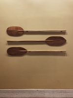 Three wooden paddles are mounted in parallel on a mustard-colored wall. Each paddle has a distinct shape and is horizontally aligned, showcasing the grain and rich brown color of the wood.