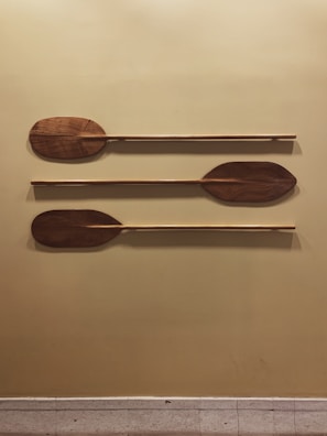 Three wooden paddles are mounted in parallel on a mustard-colored wall. Each paddle has a distinct shape and is horizontally aligned, showcasing the grain and rich brown color of the wood.