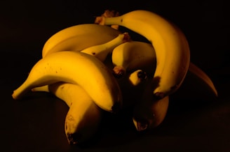 A pile of ripe bananas is arranged against a dark background, creating a strong contrast. The lighting highlights the smooth texture and bright yellow color of the bananas, with some subtle shadows adding depth.