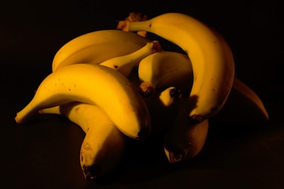 A pile of ripe bananas is arranged against a dark background, creating a strong contrast. The lighting highlights the smooth texture and bright yellow color of the bananas, with some subtle shadows adding depth.