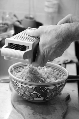 Hand using a manual grater to shred fresh cheese over a wooden cutting board.