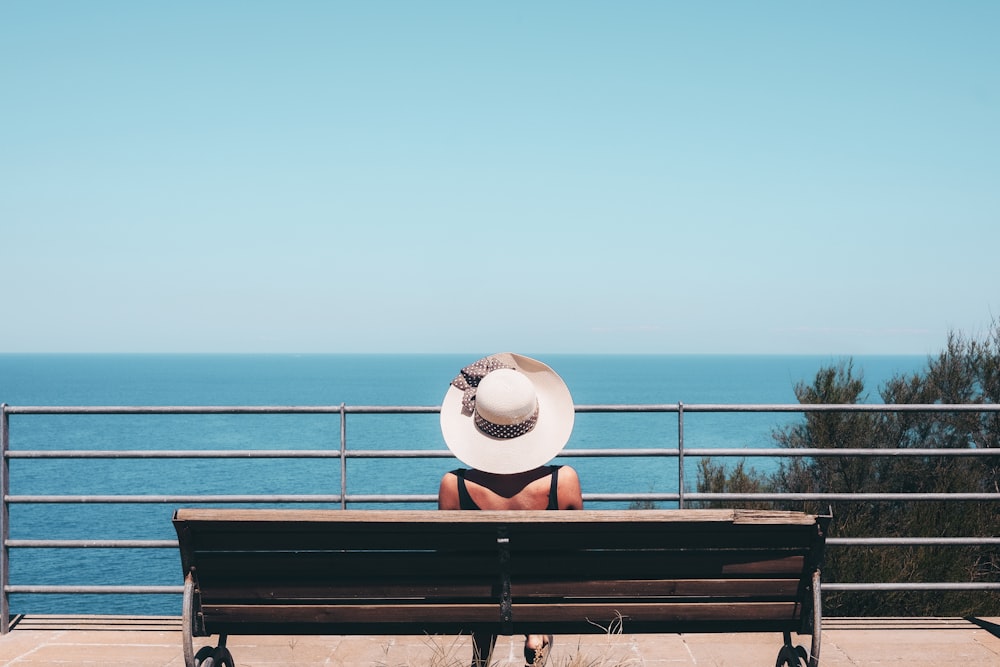 woman wearing white straw hat sitting on bench looking at sea