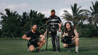 Three people are standing on a grassy field with lush green trees and palm trees in the background, holding film equipment. One person is holding a clapperboard and two others are holding cameras.