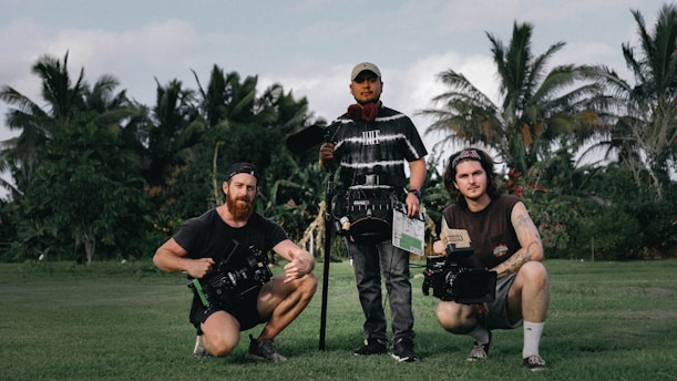 Three people are standing on a grassy field with lush green trees and palm trees in the background, holding film equipment. One person is holding a clapperboard and two others are holding cameras.