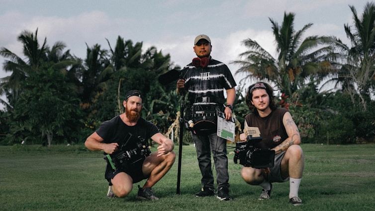 Three people are standing on a grassy field with lush green trees and palm trees in the background, holding film equipment. One person is holding a clapperboard and two others are holding cameras.