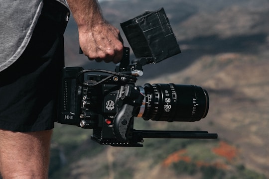 A person holding a professional camera with a large lens, standing outdoors. The camera has a matte box and various controls and dials visible on its side. The background is blurred, suggesting a natural landscape setting.