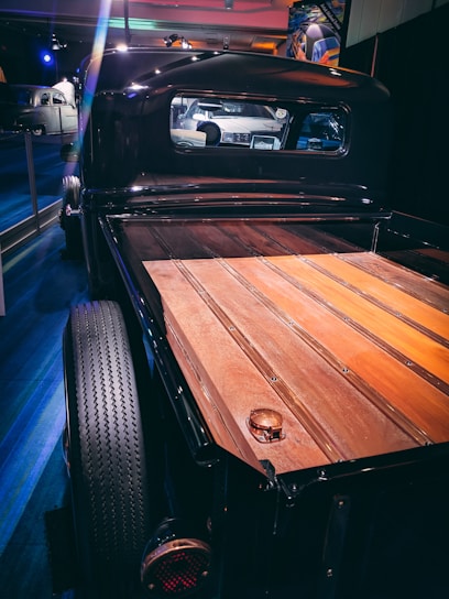 A vintage truck with a polished wooden bed is photographed indoors. The truck's black exterior shines under the modern lighting, and a classic car is visible through the rear window in the reflection. The setting appears to be a museum or exhibition space with other vehicles in the background.