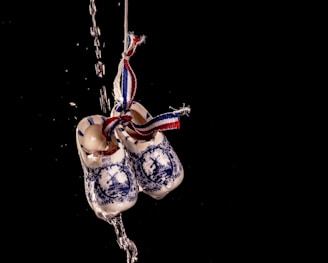 A pair of traditional ceramic Dutch clogs with windmill designs, adorned with a ribbon in blue, white, and red. The clogs are suspended in mid-air with water droplets cascading over them against a black background.