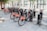 A row of rental bicycles is neatly parked at a docking station on a city street. Each bike is branded with the Santander logo in red, and the docking area is surrounded by paving stones and some trees. A red telephone booth is visible in the background, adding a classic urban element to the scene.
