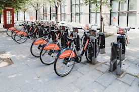 A row of rental bicycles is neatly parked at a docking station on a city street. Each bike is branded with the Santander logo in red, and the docking area is surrounded by paving stones and some trees. A red telephone booth is visible in the background, adding a classic urban element to the scene.