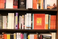 A bookshelf filled with various books of different sizes and colors. The books are neatly arranged in multiple rows. In the center of the middle shelf, there is a prominent book titled 'American Like Me' with a bright orange cover and yellow text.