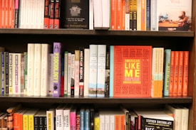 A bookshelf filled with various books of different sizes and colors. The books are neatly arranged in multiple rows. In the center of the middle shelf, there is a prominent book titled 'American Like Me' with a bright orange cover and yellow text.