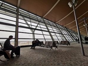 A spacious airport terminal with high ceilings and large glass windows overlooks the runway. Several people sit on metal and leather seats, looking out at the parked airplanes and the overcast sky. The architecture features a sleek and modern design with horizontal and vertical lines.