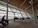A spacious airport terminal with high ceilings and large glass windows overlooks the runway. Several people sit on metal and leather seats, looking out at the parked airplanes and the overcast sky. The architecture features a sleek and modern design with horizontal and vertical lines.