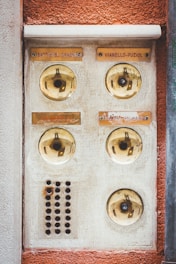An old, weathered intercom system with four round buzzer buttons, each labeled with different names. The surface shows signs of wear and aging, with a rusty grill section below the left column of buttons. The background is a mix of beige and reddish-brown textures, giving an antique look.