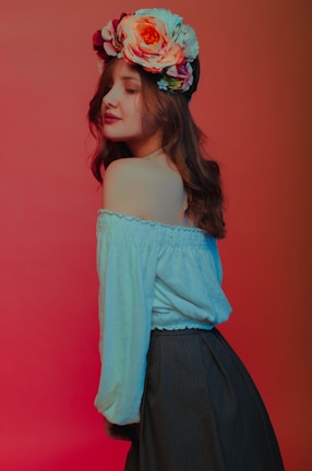 A person with long, wavy brown hair is wearing a floral crown composed of large, colorful flowers. They are dressed in an off-the-shoulder white blouse and a dark skirt, set against a gradient background of red and orange tones.