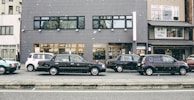 A row of parked cars is lined up along a street in front of a gray building. Snowflakes are gently falling, adding a serene atmosphere. The building features large windows and contains a sign for kimono rental services. Various cars, including a black sedan and a white compact car, are visible.