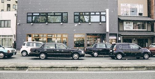 A row of parked cars is lined up along a street in front of a gray building. Snowflakes are gently falling, adding a serene atmosphere. The building features large windows and contains a sign for kimono rental services. Various cars, including a black sedan and a white compact car, are visible.
