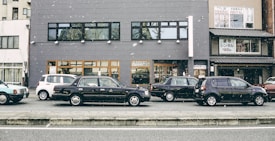 A row of parked cars is lined up along a street in front of a gray building. Snowflakes are gently falling, adding a serene atmosphere. The building features large windows and contains a sign for kimono rental services. Various cars, including a black sedan and a white compact car, are visible.