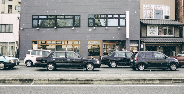 A row of parked cars is lined up along a street in front of a gray building. Snowflakes are gently falling, adding a serene atmosphere. The building features large windows and contains a sign for kimono rental services. Various cars, including a black sedan and a white compact car, are visible.