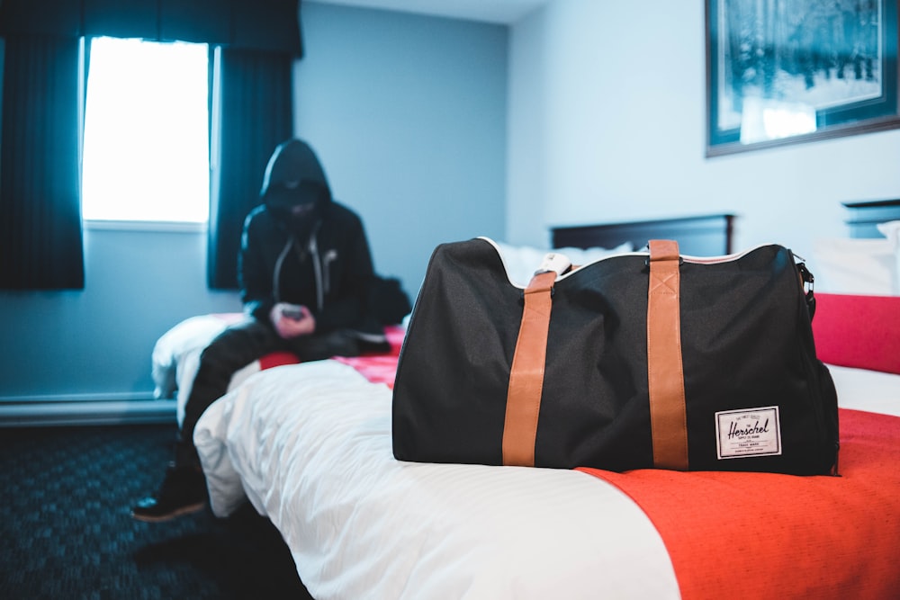 man in black hooded jacket sitting on bed near black and brown leather tote bag