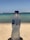 A plastic water bottle held up against a backdrop of a beach with sand and the ocean. The label on the bottle reads 'Nestlé Pure Life'. The bottle is slightly condensation-covered, suggesting a cool temperature.