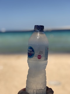 A plastic water bottle held up against a backdrop of a beach with sand and the ocean. The label on the bottle reads 'Nestlé Pure Life'. The bottle is slightly condensation-covered, suggesting a cool temperature.
