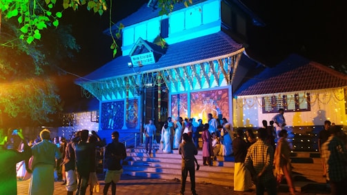 A group of people wearing traditional attire gather in front of an illuminated building decorated with lights and colorful garlands. The architecture suggests a temple or cultural site, with artwork and carvings visible on the walls. The scene is vibrant with activity under a night sky.