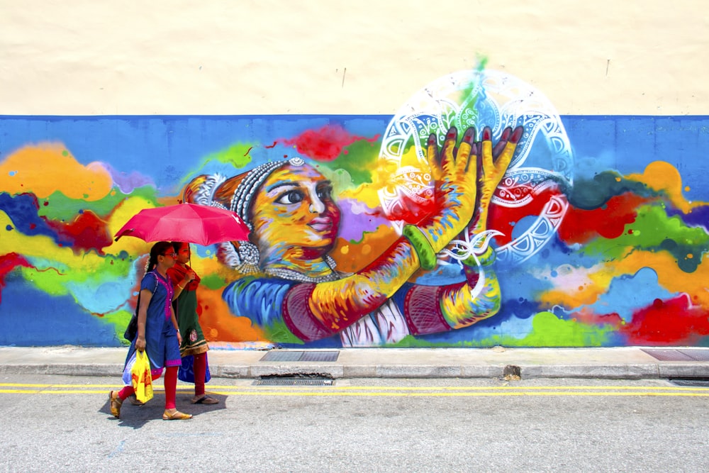 two girl holding umbrella while walking beside graffiti