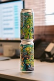 Two beverage cans with colorful and artistic designs are stacked vertically on a desk. The designs feature vibrant cartoon-style graphics, possibly depicting sea creatures or an underwater theme. The background reveals a blurred office setting with a computer screen and telephone.