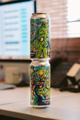 Two beverage cans with colorful and artistic designs are stacked vertically on a desk. The designs feature vibrant cartoon-style graphics, possibly depicting sea creatures or an underwater theme. The background reveals a blurred office setting with a computer screen and telephone.