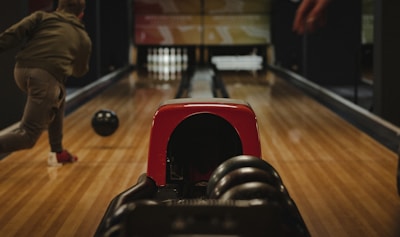 A person is bowling at a lane with wooden flooring, releasing a black bowling ball towards a set of pins. The scene captures the moment after the ball is thrown, with the person in motion wearing casual clothing and bright red shoes. The image is framed to show the lane's perspective with equipment visible in the foreground.