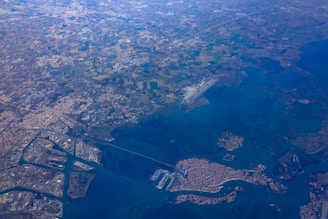 Aerial view of a coastal city with a distinct canal system, surrounded by extensive land and sea areas. The landscape shows a combination of urban structures, agricultural fields, and waterways, with a large airport visible near the center.