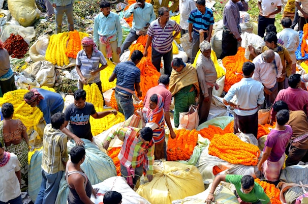 A bustling flower market with numerous vendors and buyers interacting amidst large bundles of vibrant orange and yellow marigold garlands. The scene is lively and crowded, with people standing close to each other, some negotiating prices, and others handling the flowers. The flowers are arranged in large, colorful mounds, creating a vivid display.