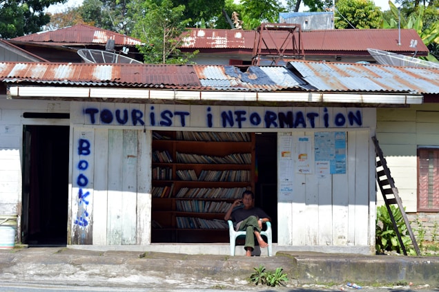 A small, rustic tourist information center with corrugated metal roofing and weathered wooden walls. Shelves filled with books are visible through the open front. A man is sitting in a chair outside, appearing relaxed. The words 'Tourist Information' and 'Books' are painted in blue on the building.