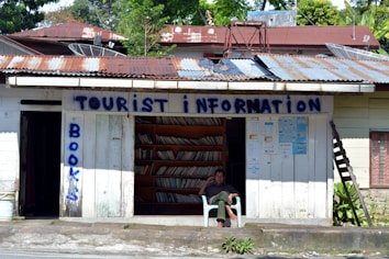 A small, rustic tourist information center with corrugated metal roofing and weathered wooden walls. Shelves filled with books are visible through the open front. A man is sitting in a chair outside, appearing relaxed. The words 'Tourist Information' and 'Books' are painted in blue on the building.