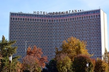The image features a large hotel building with a grid-like facade, identified by the sign on top reading 'Hotel Uzbekistan'. Surrounding the structure are trees with foliage in autumn colors, adding a natural element to the urban scene.