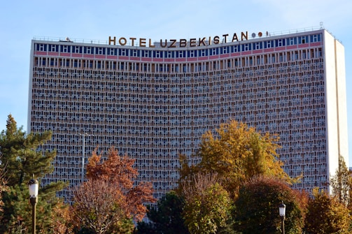 The image features a large hotel building with a grid-like facade, identified by the sign on top reading 'Hotel Uzbekistan'. Surrounding the structure are trees with foliage in autumn colors, adding a natural element to the urban scene.