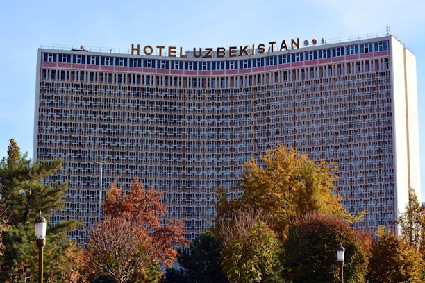 The image features a large hotel building with a grid-like facade, identified by the sign on top reading 'Hotel Uzbekistan'. Surrounding the structure are trees with foliage in autumn colors, adding a natural element to the urban scene.