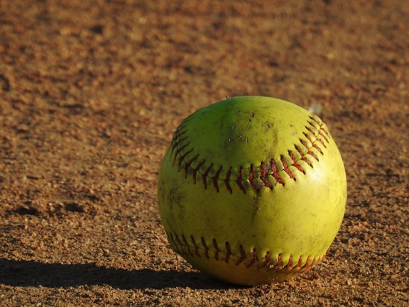 A yellow softball with red stitching is resting on a dirt field. The surface of the ball is slightly worn, indicating it may have been used in play. The lighting creates a warm tone, and the texture of the dirt is clearly visible.