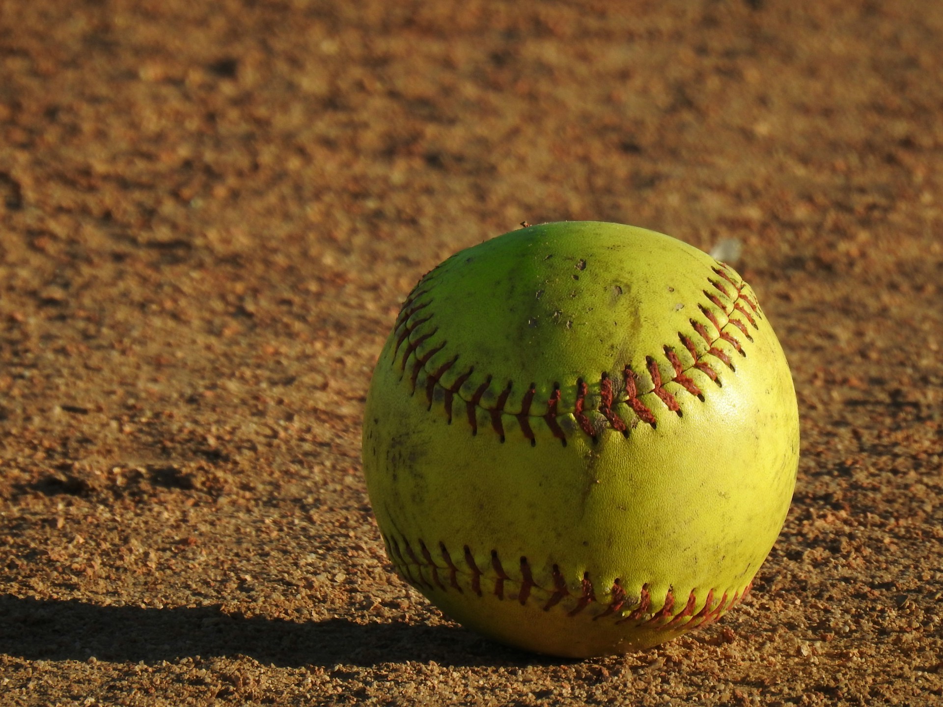 A yellow softball with red stitching is resting on a dirt field. The surface of the ball is slightly worn, indicating it may have been used in play. The lighting creates a warm tone, and the texture of the dirt is clearly visible.