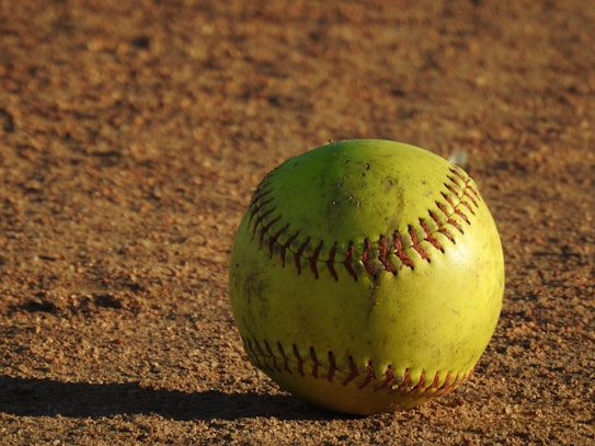 A yellow softball with red stitching is resting on a dirt field. The surface of the ball is slightly worn, indicating it may have been used in play. The lighting creates a warm tone, and the texture of the dirt is clearly visible.