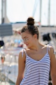 A person with glasses and a bun hairstyle is wearing a striped sleeveless top. The background suggests a marina or harbor setting with boats and masts visible.