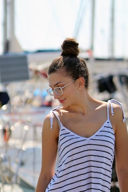 A person with glasses and a bun hairstyle is wearing a striped sleeveless top. The background suggests a marina or harbor setting with boats and masts visible.