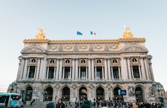 The image features an ornate neoclassical architectural building with intricate sculptures and decorations. The façade is adorned with columns and detailed carvings, while two golden statues are mounted on its top corners. Two flags, one blue and the other with a tricolor design, flutter above the structure. A crowd of people and a green bus are present in the foreground, indicating a bustling urban environment.