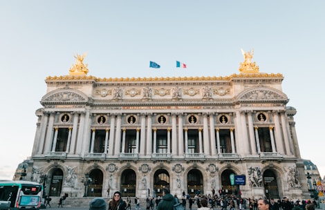 The image features an ornate neoclassical architectural building with intricate sculptures and decorations. The façade is adorned with columns and detailed carvings, while two golden statues are mounted on its top corners. Two flags, one blue and the other with a tricolor design, flutter above the structure. A crowd of people and a green bus are present in the foreground, indicating a bustling urban environment.