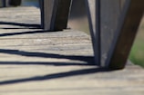 A close-up view of a wooden deck with vertical posts casting strong shadows across the surface. The perspective emphasizes the texture of the wood grain and the contrast between the light and shadow.
