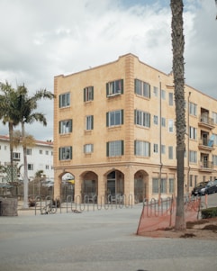A four-story, beige apartment building with numerous windows and arched entrances stands prominently. The foreground features bike racks and palm trees, with a cloudy sky overhead.