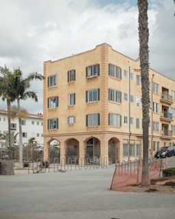 A four-story, beige apartment building with numerous windows and arched entrances stands prominently. The foreground features bike racks and palm trees, with a cloudy sky overhead.